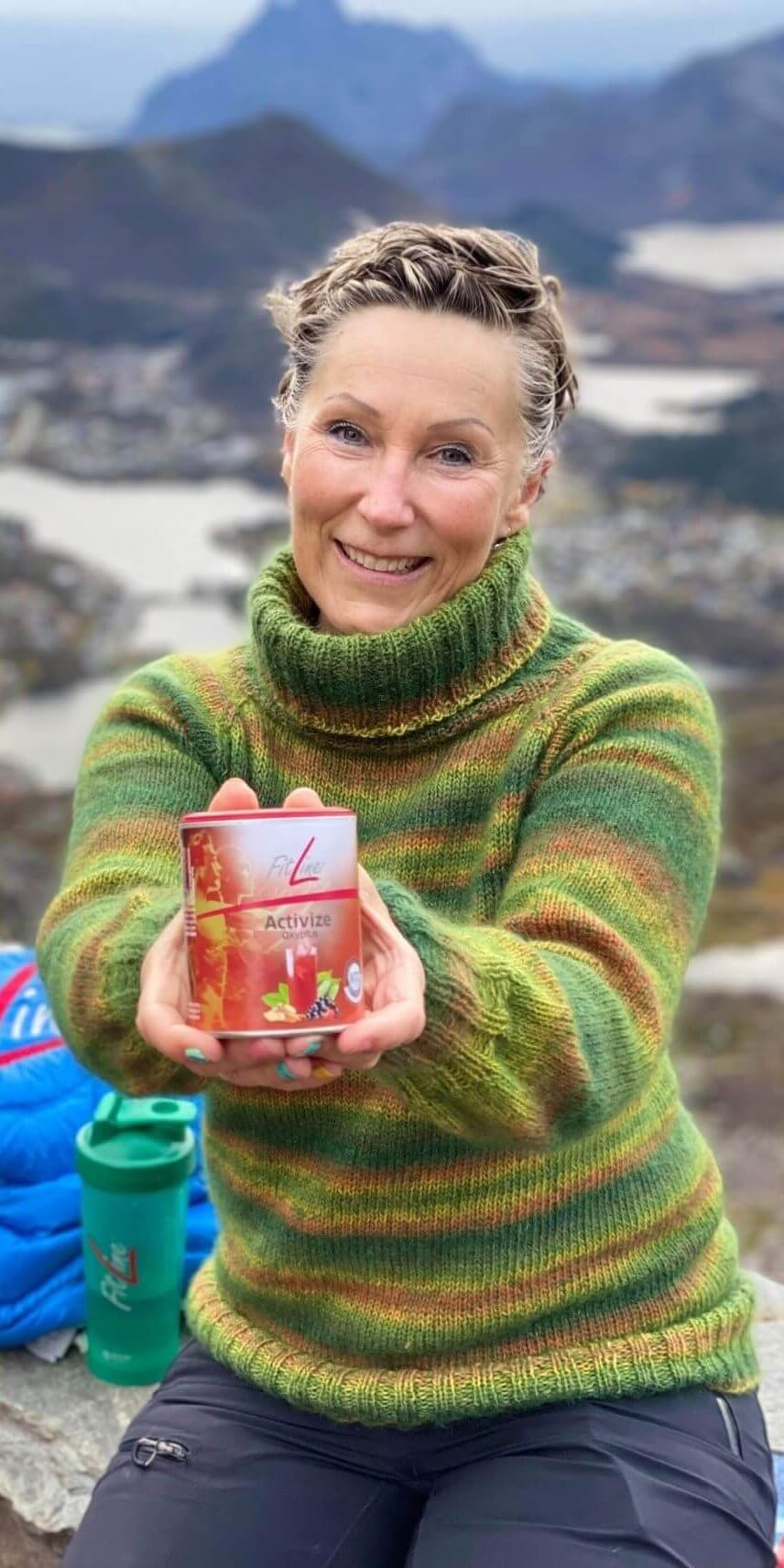 Una mujer madura con reflejos rubios sonriendo y sosteniendo un envase de FitLine Activize entre sus manos, con montañas al fondo.