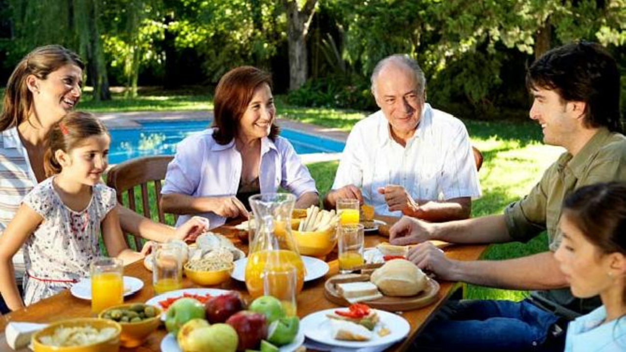 Familia de 6 personas comiendo comida sana en una mesa en el campo, rodeados de árboles y con una piscina en el fondo.